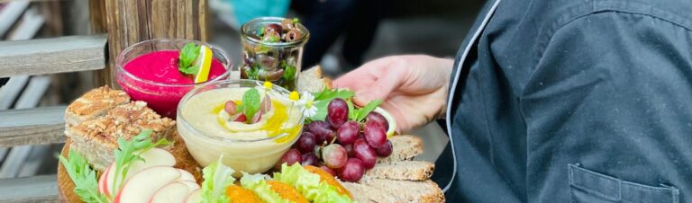 Holzplatte, die von einer Person in schwarzer Schürze gehalten wird. Darauf verschiedene Leckereien wie Trauben, Humus, Brot, Äpfel und mehr.