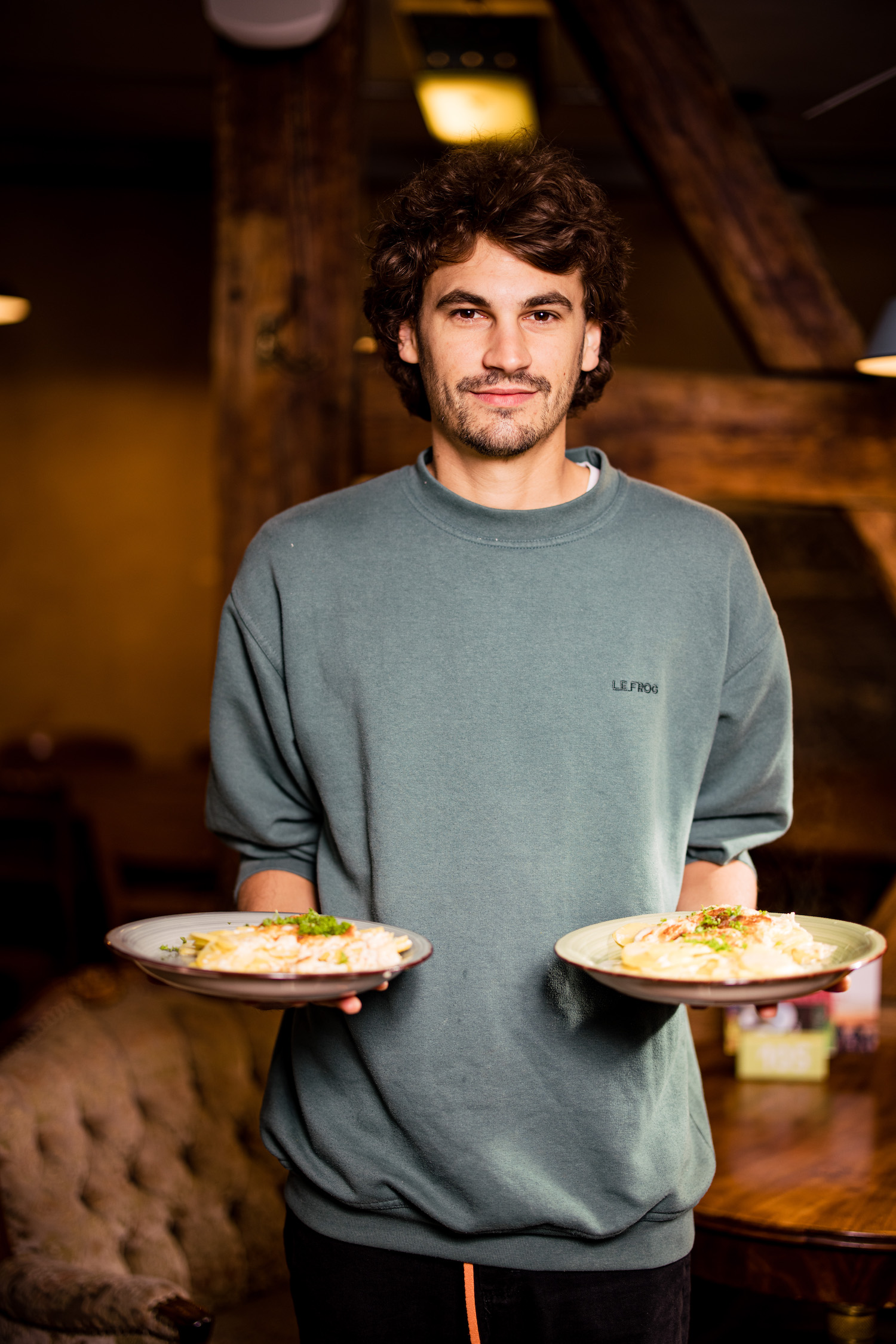 Ein Ehrenamtlicher der Kulturküche mit grauem Pullover und braunen Haaren hat zwei Teller mit Essen in der Hand.