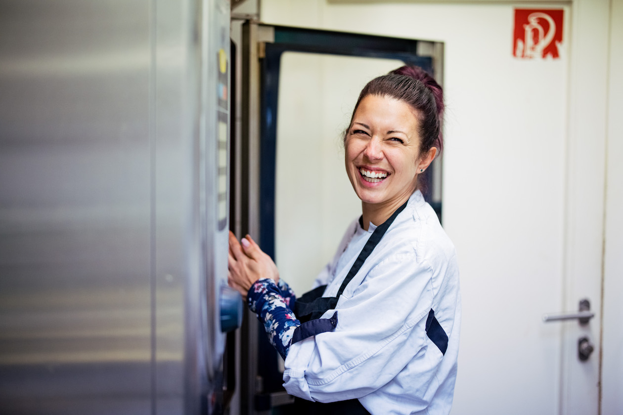 Eine Junge Frau in weißer Küchen-Schürze steht am großen Ofen und lacht in die Kamera.
