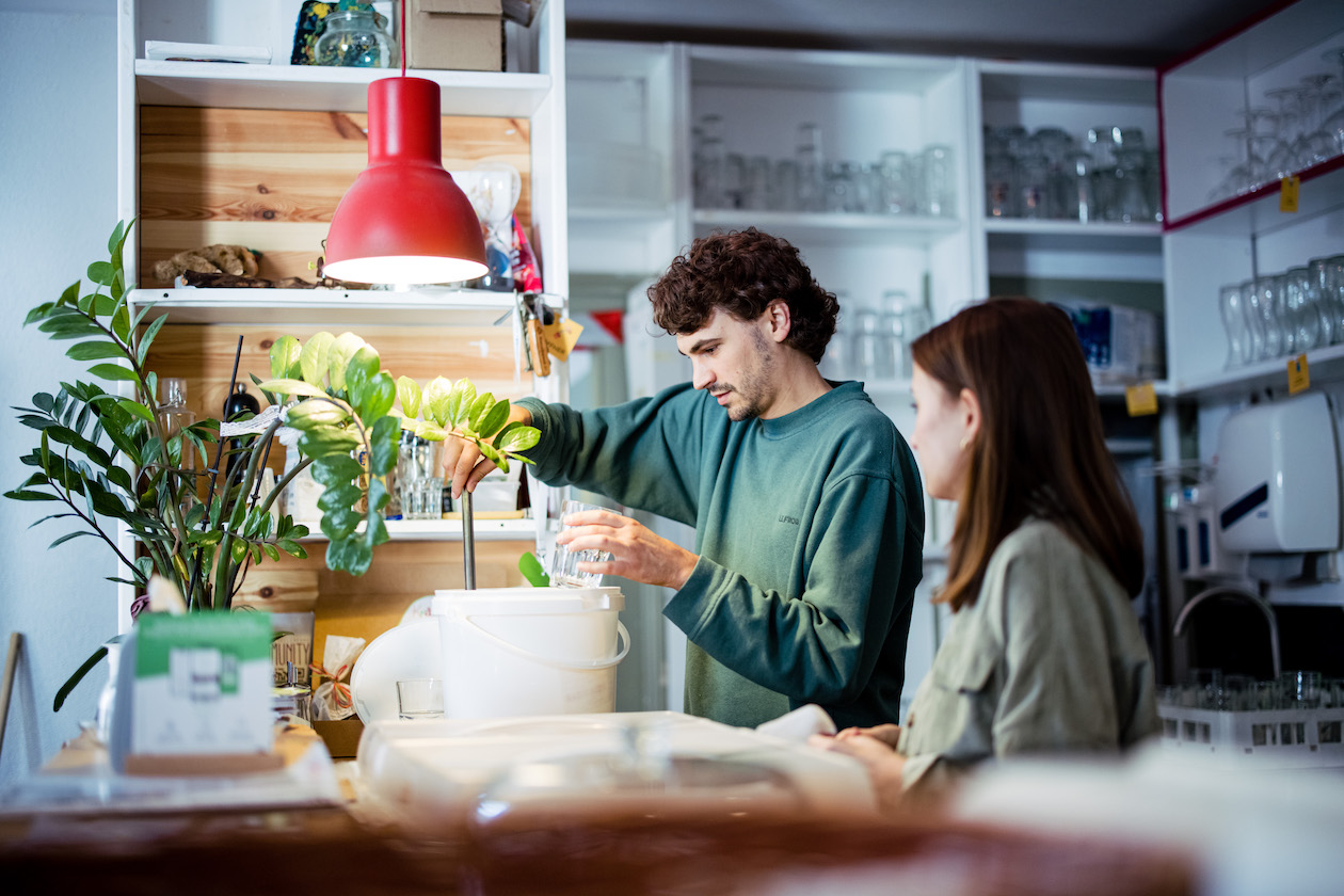 Ein Mann und eine Frau stehen hinter der Theke der Kulturküche. Über der Theke hängt eine rote Lampe und auf der Theke steht eine Goldfeder.