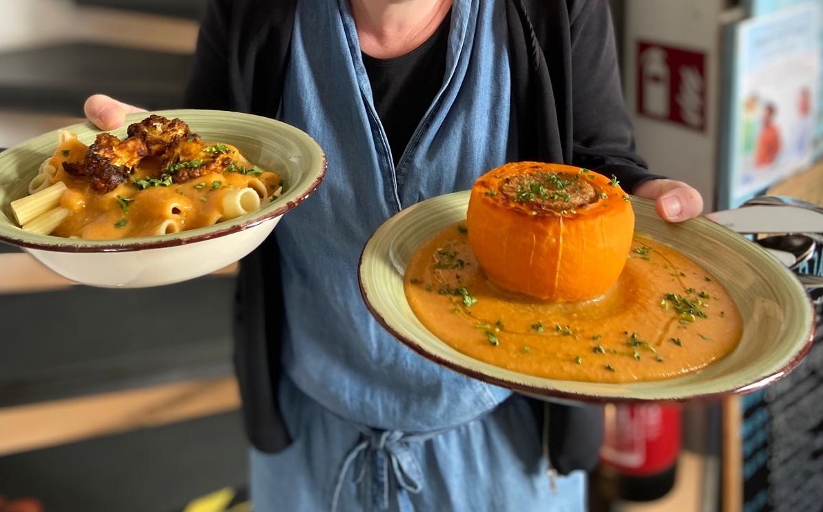 Eine Person hält zwei Teller, auf dem einen ist ein gefüllter orangener Kürbis und auf dem anderen Nudeln mit einer Soße.