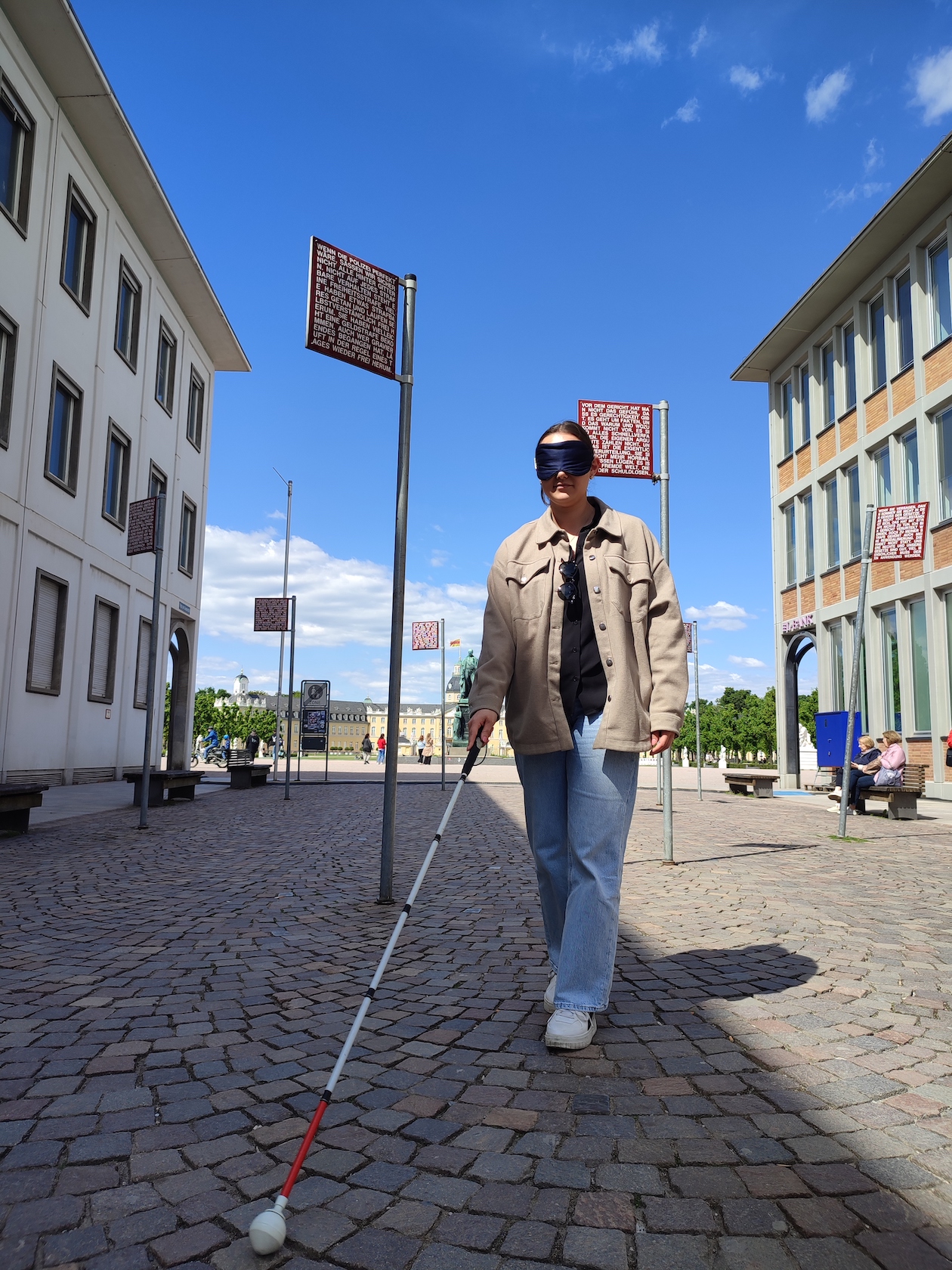 Auf dem Platz der Grundrechte geht eine Frau mit Blindenstock. Sie hat eine Augenmaske auf. Das Bild ist im Rahmen eines unserer Projekte entstanden. Menschen konnten ausprobieren, wie es sich mit weniger Sehvermögen anfühlt.