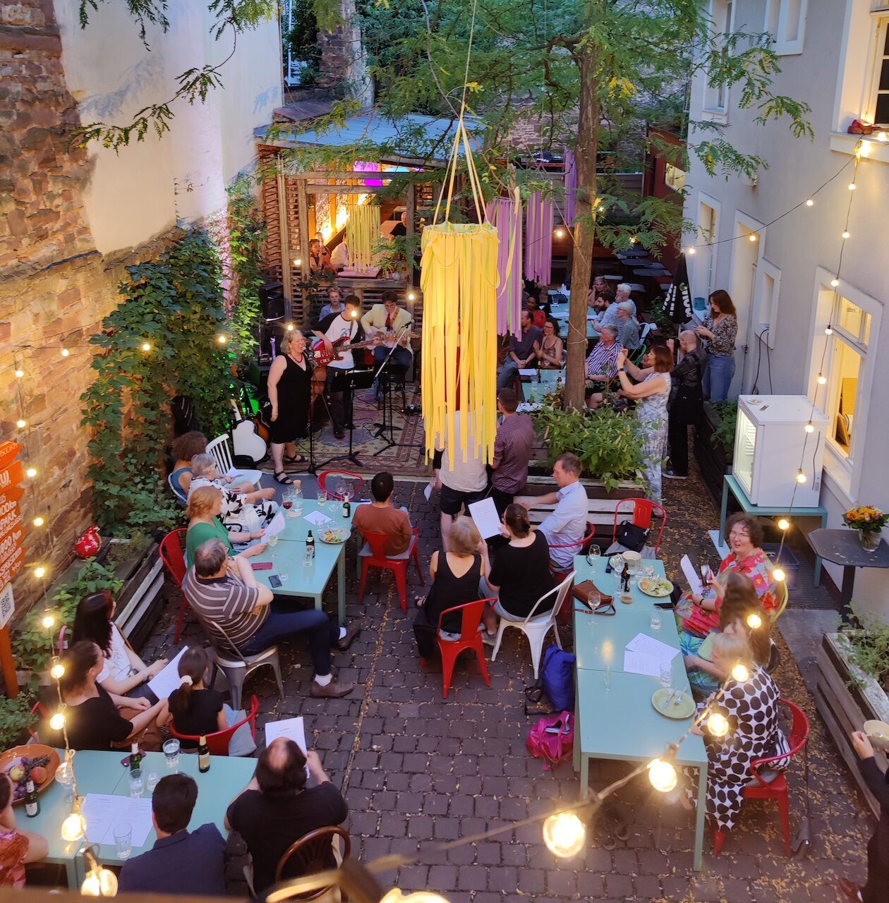 Innenhof der Kulturküche, beleuchtet mit Lichterketten und bunte Girlanden. Auf einem Teppich stehen Musikerinnen und Musiker. Im Hof sitzen viele Menschen auf Stühlen oder stehen.