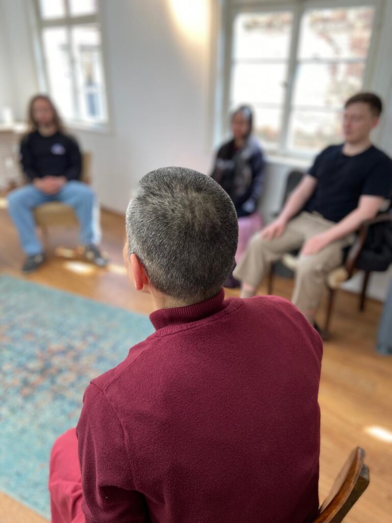 Vier Personen sitzen in einem Raum auf Stühlen mit geschlossenen Augen