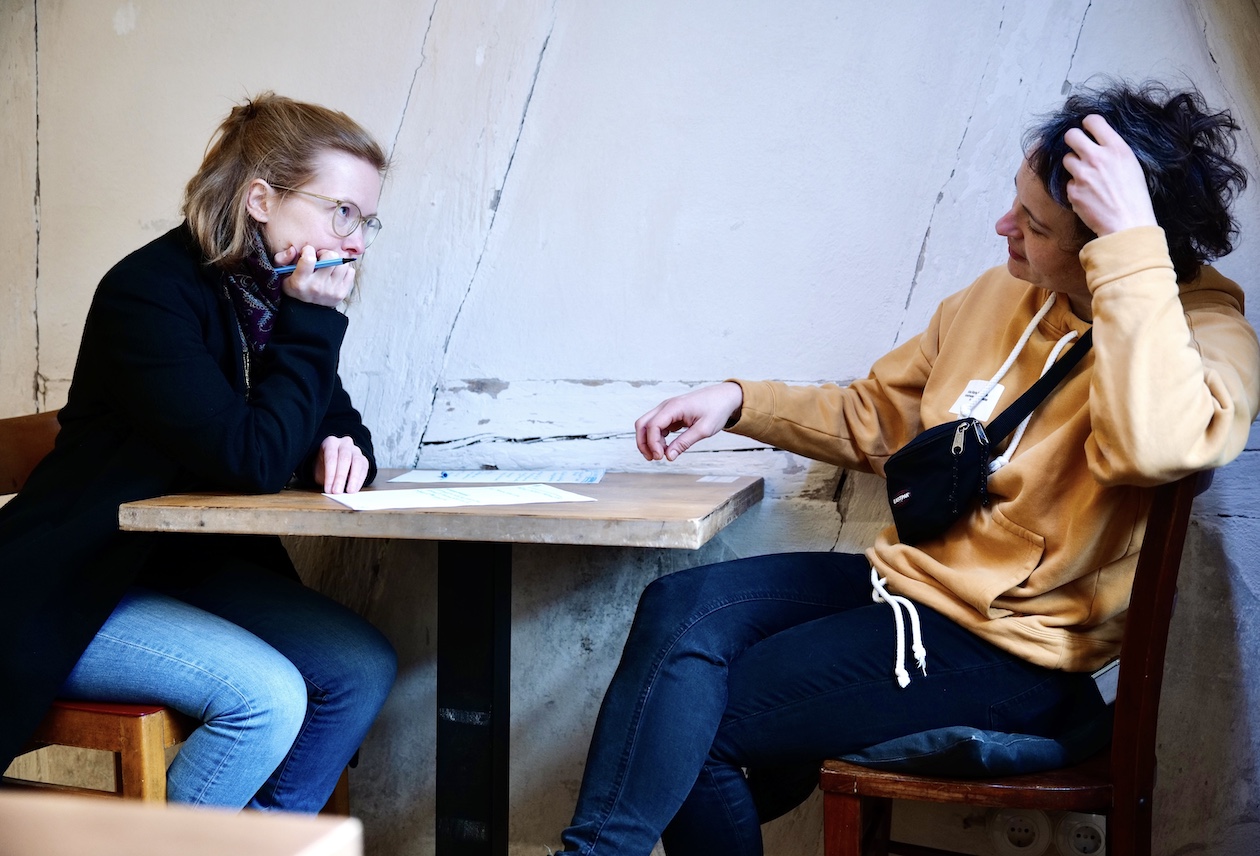 Zwei Frauen sitzen an einem Tisch und besprechen sich. Die eine hat einen Stift in der Hand und stürzt sich ab. Auf dem Tisch liegen Zettel.