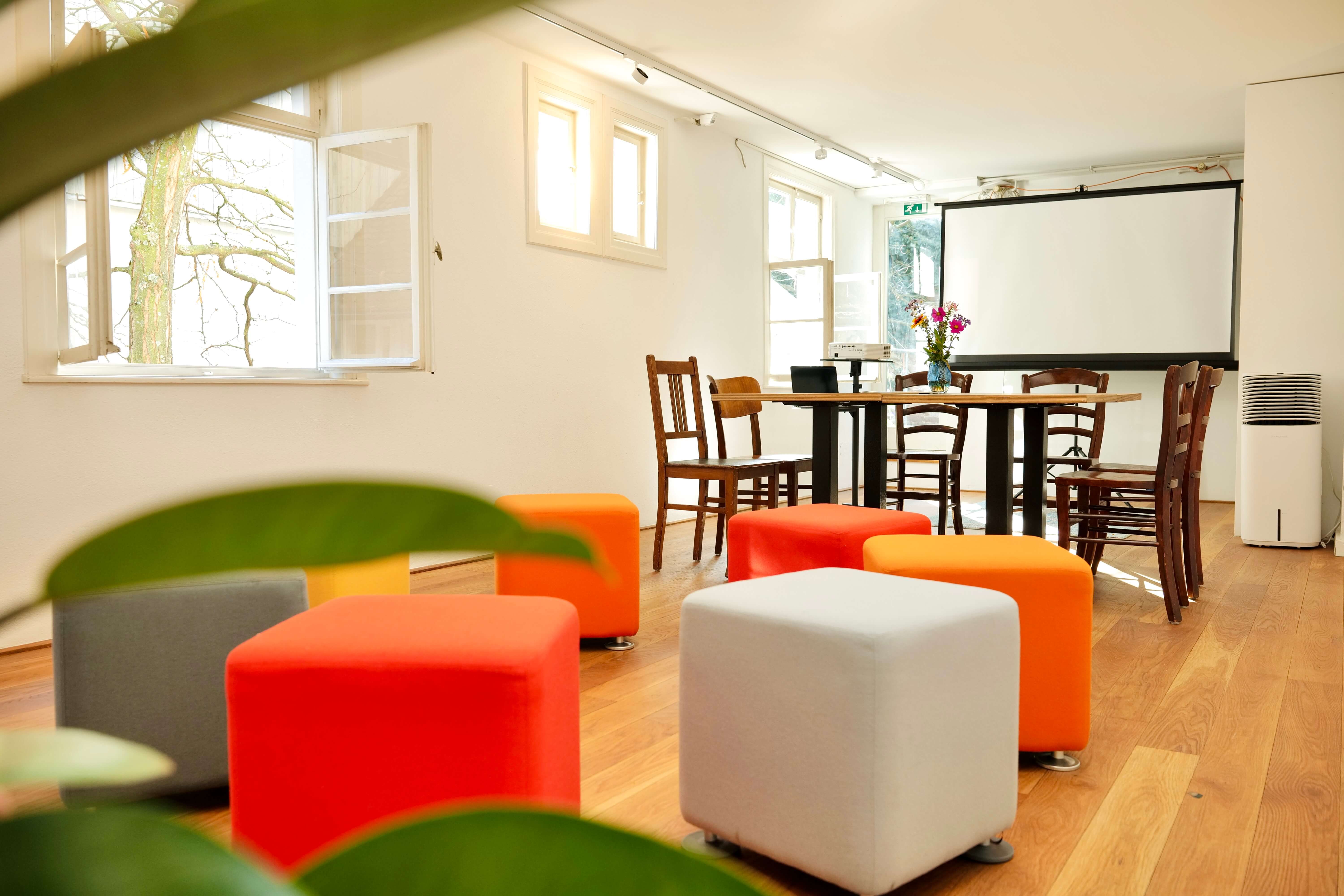 Heller Seminarraum, in dem Würfehocker in weiß und orange stehen. Im Hintergrund sieht man eine Leinwand. 