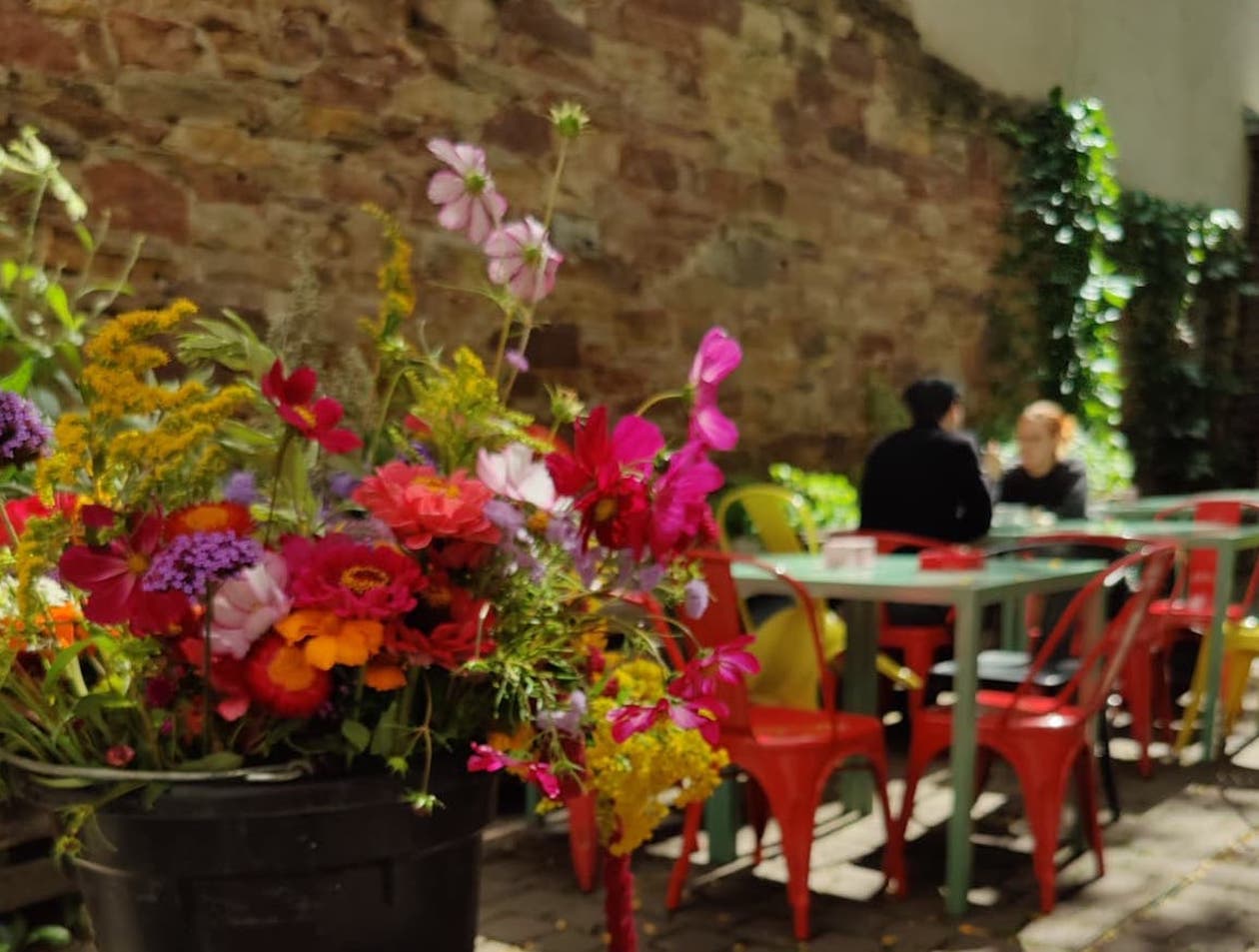 Bild von einem bunten Blumenstrauß. Im Hintergrund der Innenhof der Kulturküche mit den grünen Tischen und bunten Stühlen.