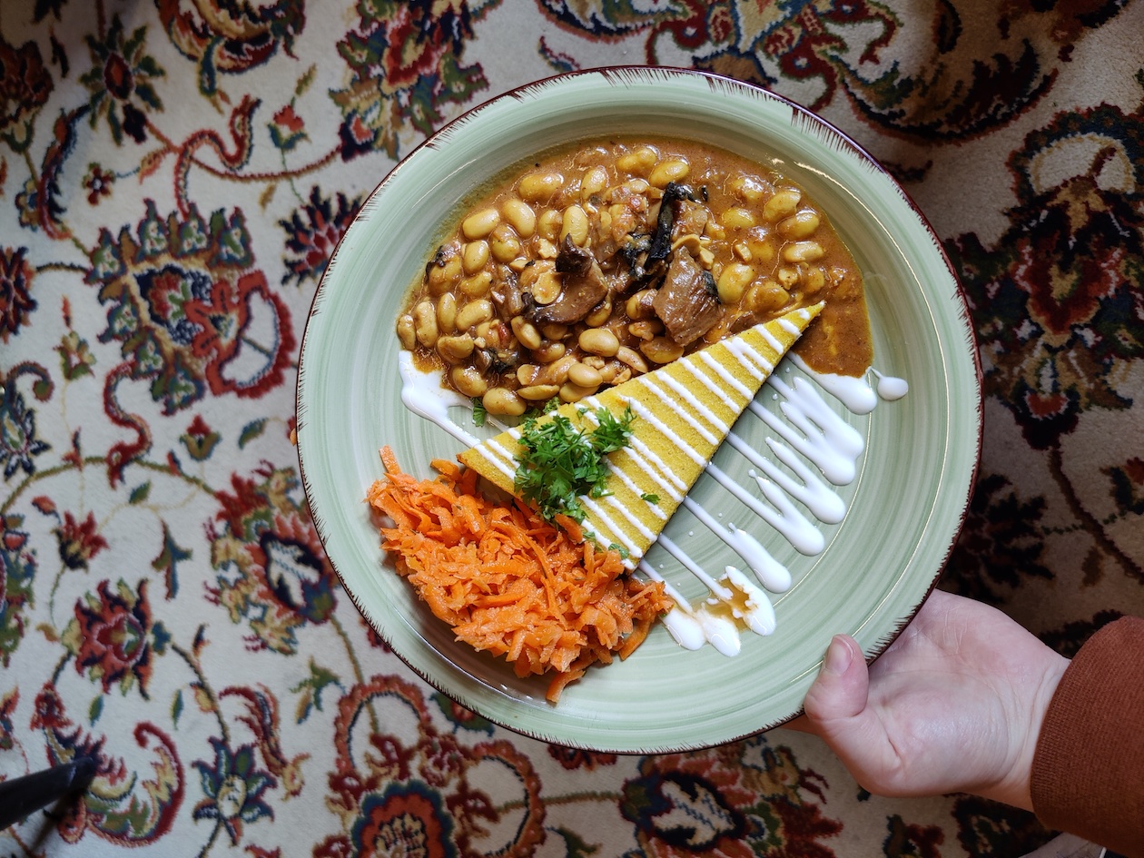 Bunter Teppich über den eine Person einen Teller mit Essen hält. Auf dem Teller ist ein Gericht mit Karottensalat, Polenta-Schnitte, Bohnen und Topping.