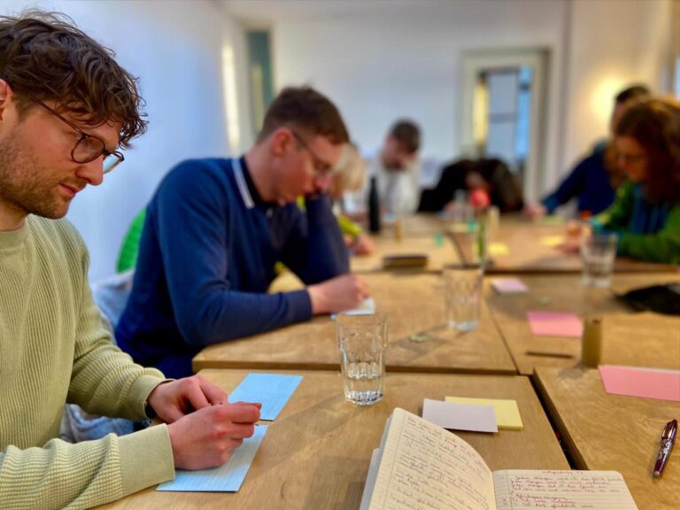 Personen sitzen an einem Tisch und schreiben Gedanken auf Zettel.