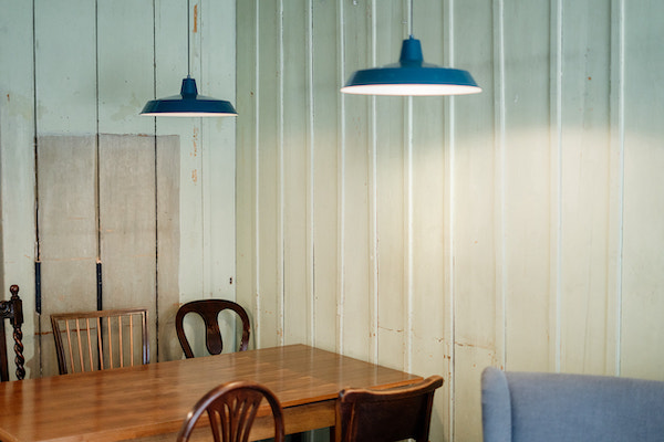 Raum mit Wandvertäfelung. Von der Decke hängen zwei blaue Lampen. Außerdem ein Holztisch mit fünf Holzstühlen.