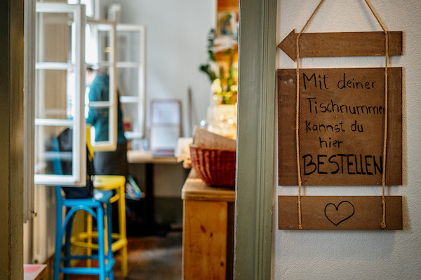 Auf einem Holzschild steht "Mit deiner Tischnummer kannst du hier bestellen". Daneben ist ein Türrahmen, im Hintergrund sind offene Fenster und zwei bunte Stühle zu sehen.
