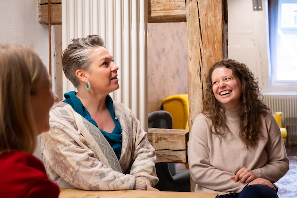 Zwei Frauen sitzen nebeneinander. Im Hintergrund sieht man Holzbalken des alten Hauses.