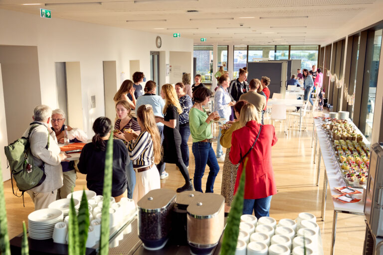 großer Raum mit Menschen, die sich unterhalten. Rechts im Bild ein Buffet.